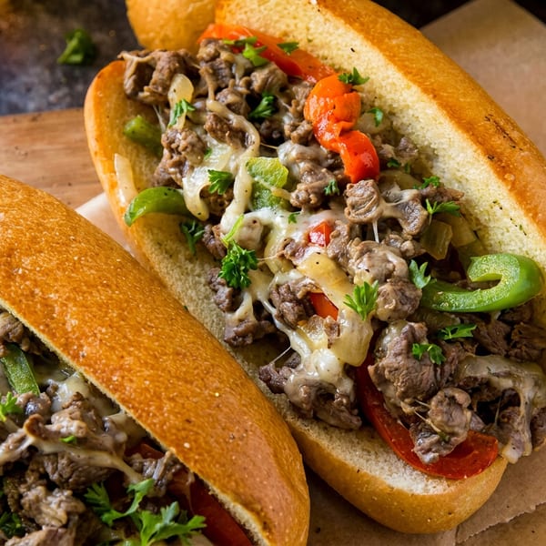Two delicious cheesesteak sandwiches with melted cheese, peppers, and onions on a wooden board.